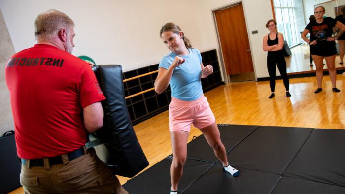 A student with raised fists prepares to demonstrate a punching technique. An instructor stands holding a thick safety pad to absorb the impact. Two students watch a few feet away.